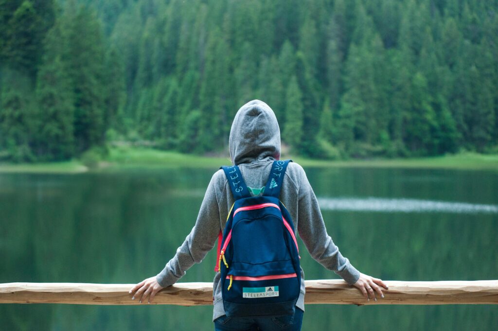 A person in a hoodie stands by a serene lake surrounded by lush greenery, enjoying the peaceful outdoors.