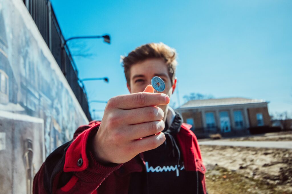 A young adult holding a coin outdoors with a clear blue sky and building in the background.