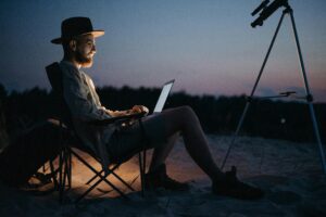 A man sitting on a beach with a laptop and telescope, enjoying the sunset.