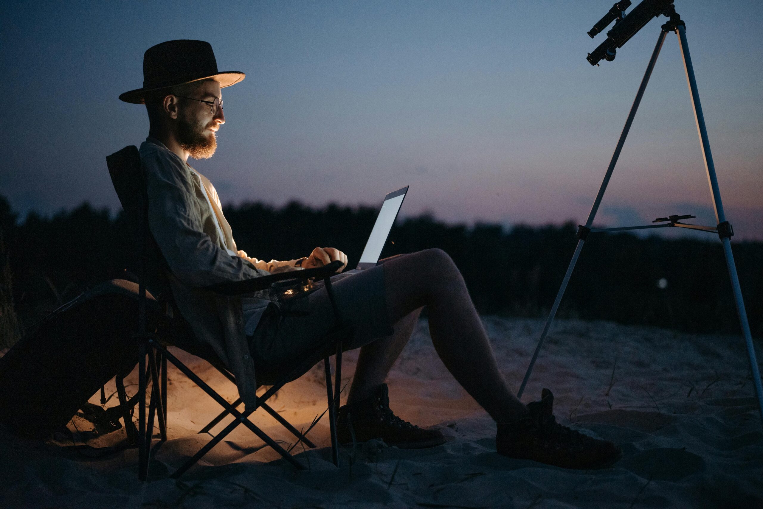 A man sitting on a beach with a laptop and telescope, enjoying the sunset.