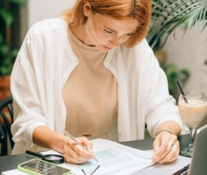 Woman analyzing financial documents using laptop and calculator indoors.